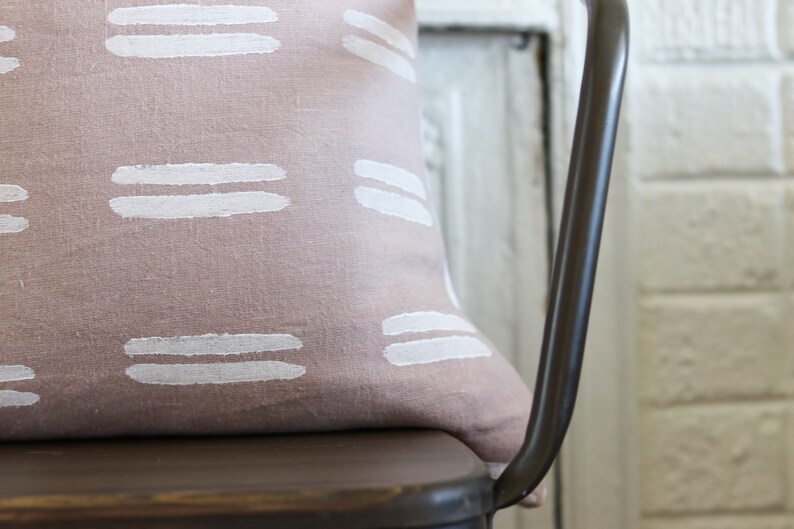 May include: A decorative pillow with a dusty rose background and white horizontal brushstroke patterns. The pillow is resting on a dark brown metal chair. The background includes a white brick wall and a white door.