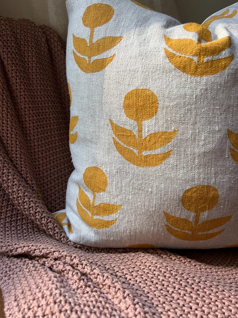 Puede incluir: Un coj&iacute;n decorativo con un fondo gris claro y un patr&oacute;n repetido de dise&ntilde;os florales amarillos. El coj&iacute;n descansa sobre una manta de punto rosa. El dise&ntilde;o floral presenta un centro circular y tres hojas curvas.