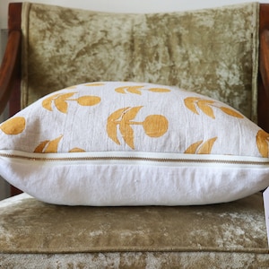 Puede incluir: Un coj&iacute;n decorativo con una base blanca y un patr&oacute;n de dise&ntilde;os florales amarillos. El coj&iacute;n tiene una cremallera dorada y una etiqueta que dice "MUSE textiles hand printed home goods". El coj&iacute;n est&aacute; sobre una silla vintage.