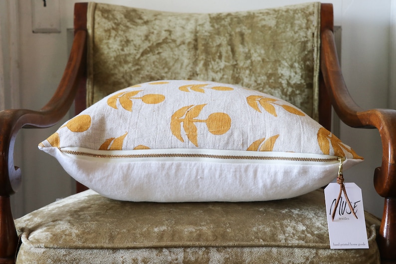 Puede incluir: Un coj&iacute;n decorativo con una base blanca y un patr&oacute;n de dise&ntilde;os florales amarillos. El coj&iacute;n tiene una cremallera dorada y una etiqueta que dice "MUSE textiles hand printed home goods". El coj&iacute;n est&aacute; sobre una silla vintage.