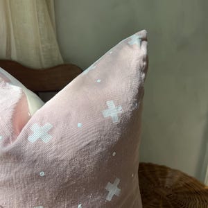 Może przedstawiać: Zbliżenie r&oacute;żowej poduszki z białym wzorem krzyża. Poduszka wykonana jest z miękkiej, teksturowanej tkaniny. Poduszka leży na drewnianym krześle.