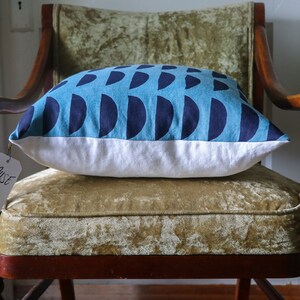 May include: A blue and white throw pillow with a geometric pattern of navy blue half circles on a light blue background. The pillow is sitting on a green velvet chair with wooden arms. A white tag with the word "Muse" is attached to the chair.