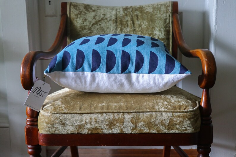 May include: A blue and white throw pillow with a geometric pattern of navy blue half circles on a light blue background. The pillow is sitting on a green velvet chair with wooden arms. A white tag with the word "Muse" is attached to the chair.
