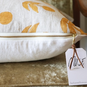Puede incluir: Un coj&iacute;n rectangular con base blanca y cremallera dorada. La tela superior presenta un patr&oacute;n repetido de dise&ntilde;os de hojas y tallos dorados. Se adjunta una etiqueta con el texto "MUSE textiles".
