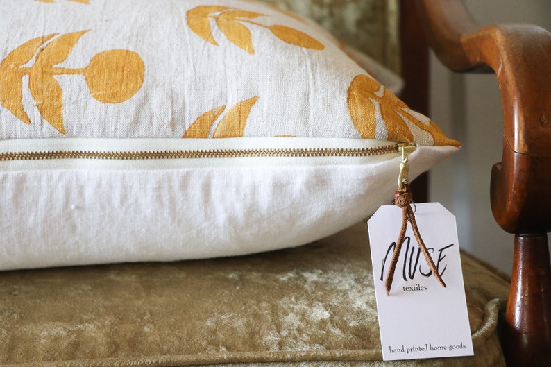Puede incluir: Un coj&iacute;n rectangular con base blanca y cremallera dorada. La tela superior presenta un patr&oacute;n repetido de dise&ntilde;os de hojas y tallos dorados. Se adjunta una etiqueta con el texto "MUSE textiles".