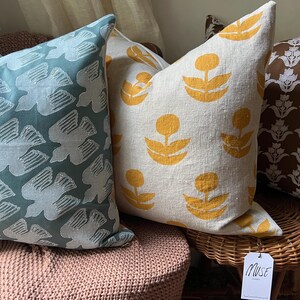 Puede incluir: Una colecci&oacute;n de cojines decorativos con varios dise&ntilde;os. Un coj&iacute;n tiene un fondo verde azulado con un dise&ntilde;o de p&aacute;jaro blanco. Otro tiene un fondo crema con formas florales amarillas. Un tercer coj&iacute;n tiene un fondo marr&oacute;n con un estampado floral blanco.