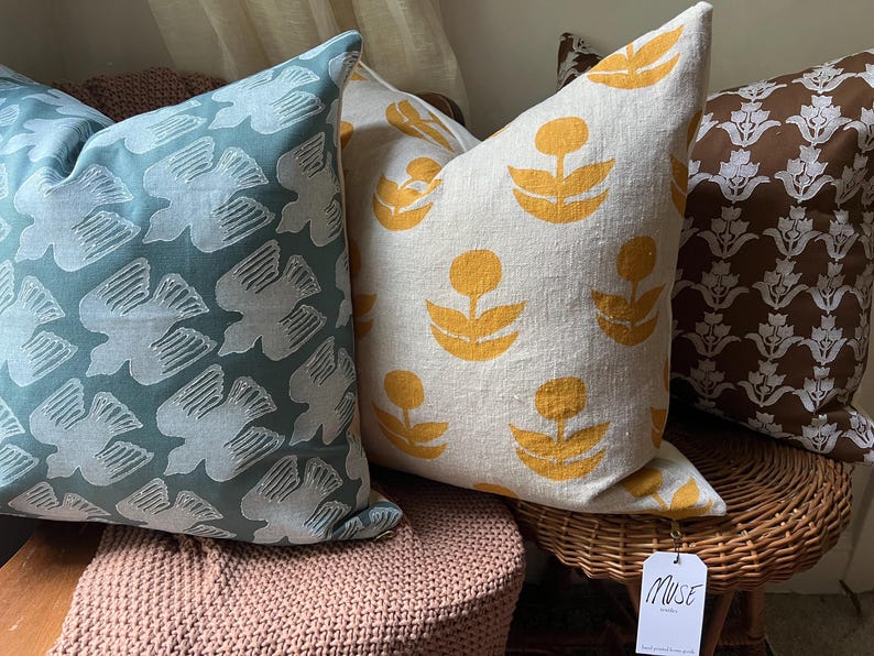 Puede incluir: Una colecci&oacute;n de cojines decorativos con varios dise&ntilde;os. Un coj&iacute;n tiene un fondo verde azulado con un dise&ntilde;o de p&aacute;jaro blanco. Otro tiene un fondo crema con formas florales amarillas. Un tercer coj&iacute;n tiene un fondo marr&oacute;n con un estampado floral blanco.