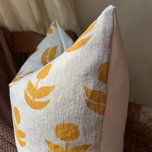 Puede incluir: Primer plano de un coj&iacute;n decorativo con tejido de lino color crema. El coj&iacute;n presenta un patr&oacute;n repetido de dise&ntilde;os florales amarillos. El lateral del coj&iacute;n es de tela blanca lisa. El coj&iacute;n es un art&iacute;culo de decoraci&oacute;n del hogar.