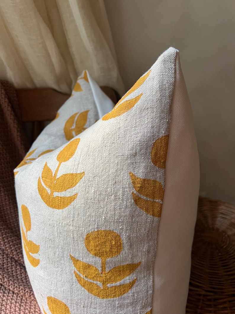 Puede incluir: Primer plano de un coj&iacute;n decorativo con tejido de lino color crema. El coj&iacute;n presenta un patr&oacute;n repetido de dise&ntilde;os florales amarillos. El lateral del coj&iacute;n es de tela blanca lisa. El coj&iacute;n es un art&iacute;culo de decoraci&oacute;n del hogar.
