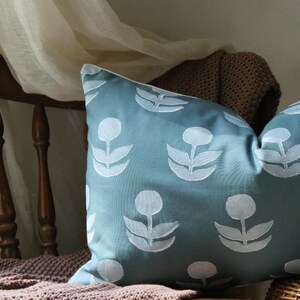 May include: A rectangular teal pillow with a white floral pattern. The pillow is resting on a brown knitted blanket and a wooden chair. The pillow's design features stylized white flowers.