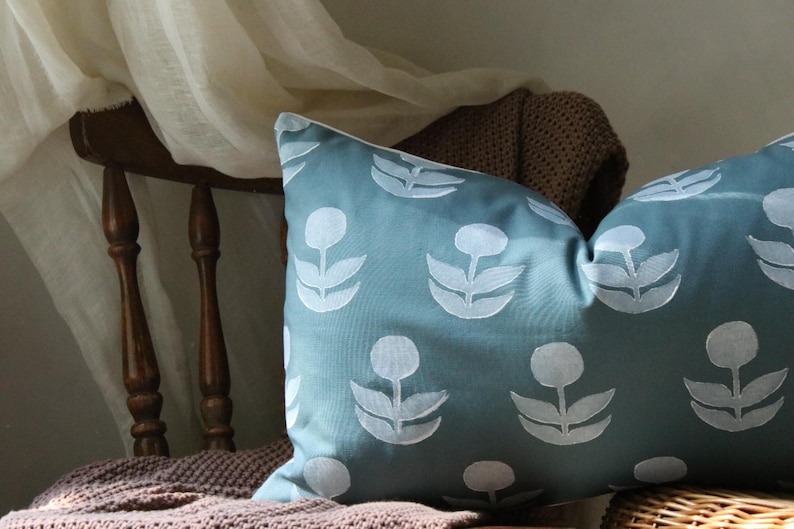 May include: A rectangular teal pillow with a white floral pattern. The pillow is resting on a brown knitted blanket and a wooden chair. The pillow's design features stylized white flowers.