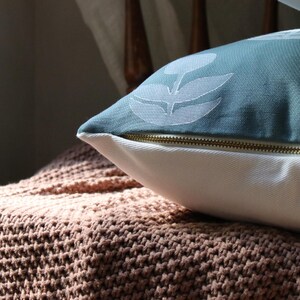 May include: A decorative pillow with a teal floral pattern and a white base, resting on a knitted throw. The pillow has a gold zipper. The throw is a light brown color.