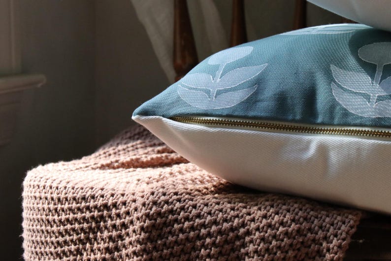 May include: A decorative pillow with a teal floral pattern and a white base, resting on a knitted throw. The pillow has a gold zipper. The throw is a light brown color.