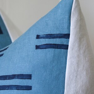 Puede incluir: Una almohada de lino azul con un borde blanco y rayas horizontales azul oscuro. La almohada est&aacute; hecha de una tela suave y texturizada.