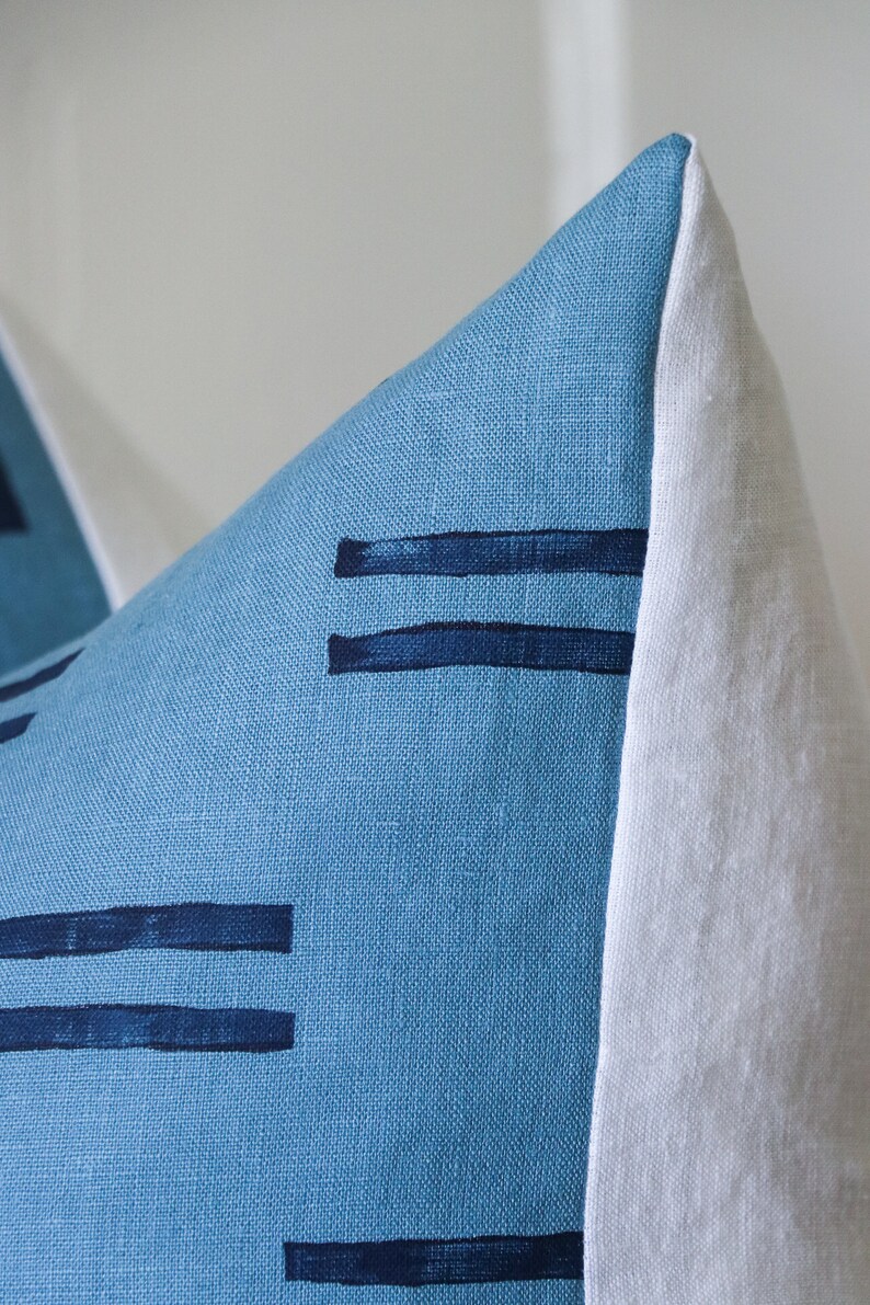 Puede incluir: Una almohada de lino azul con un borde blanco y rayas horizontales azul oscuro. La almohada est&aacute; hecha de una tela suave y texturizada.