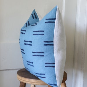 Puede incluir: Una almohada azul y blanca con un patr&oacute;n geom&eacute;trico de rayas azul oscuro. La almohada est&aacute; sentada en un taburete de madera.