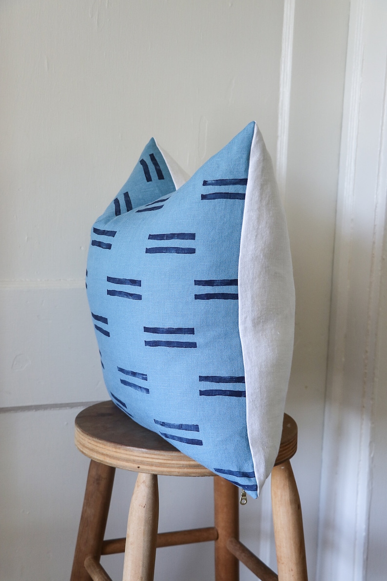 Puede incluir: Una almohada azul y blanca con un patr&oacute;n geom&eacute;trico de rayas azul oscuro. La almohada est&aacute; sentada en un taburete de madera.