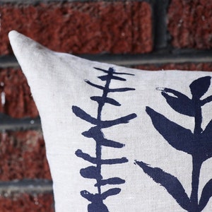 Pu&ograve; includere: Un cuscino in lino con un design botanico. Il cuscino presenta uno sfondo color crema con un motivo ripetuto di illustrazioni di piante stilizzate blu scuro. Il cuscino &egrave; posto davanti a un muro di mattoni rossi.