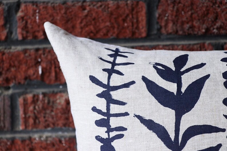 Pu&ograve; includere: Un cuscino in lino con un design botanico. Il cuscino presenta uno sfondo color crema con un motivo ripetuto di illustrazioni di piante stilizzate blu scuro. Il cuscino &egrave; posto davanti a un muro di mattoni rossi.