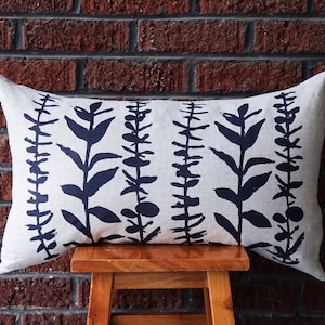 Pu&ograve; includere: Un cuscino decorativo rettangolare con sfondo color crema e un motivo ripetuto di illustrazioni botaniche blu scuro. Il cuscino &egrave; appoggiato su uno sgabello di legno davanti a un muro di mattoni.