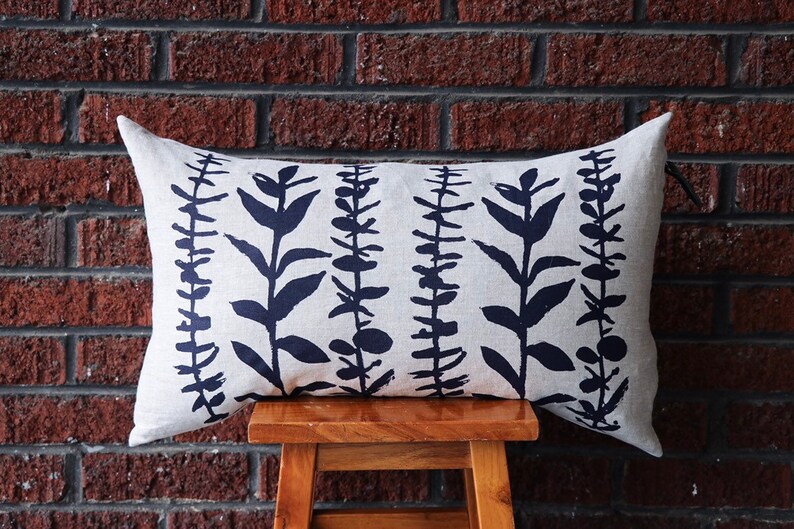 Pu&ograve; includere: Un cuscino decorativo rettangolare con sfondo color crema e un motivo ripetuto di illustrazioni botaniche blu scuro. Il cuscino &egrave; appoggiato su uno sgabello di legno davanti a un muro di mattoni.