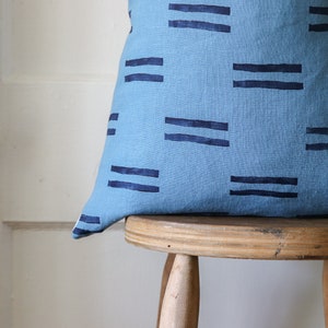 Puede incluir: Una almohada azul y azul marino con un dise&ntilde;o geom&eacute;trico. La almohada est&aacute; sentada en un taburete de madera.