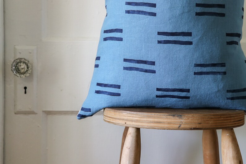 Puede incluir: Una almohada azul y azul marino con un dise&ntilde;o geom&eacute;trico. La almohada est&aacute; sentada en un taburete de madera.