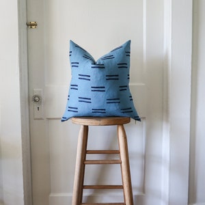 Puede incluir: Una almohada azul y negra con estampado se encuentra sobre un taburete de madera. La almohada est&aacute; hecha de una tela suave y texturizada y tiene un dise&ntilde;o geom&eacute;trico simple.