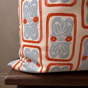 May include: A decorative pillow with a repeating pattern of light blue floral designs within orange rectangular frames on a white background. The pillow is resting on a dark wooden surface.