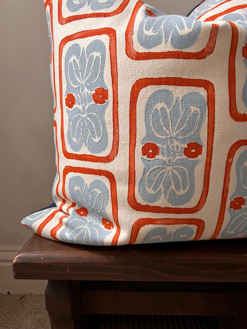 May include: A decorative pillow with a repeating pattern of light blue floral designs within orange rectangular frames on a white background. The pillow is resting on a dark wooden surface.