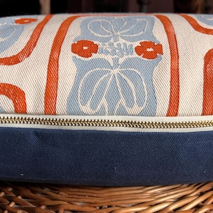 May include: Decorative pillow with a blue, white, and orange floral pattern. The pillow has a navy blue base and a brass zipper. The pillow is resting on a woven basket.