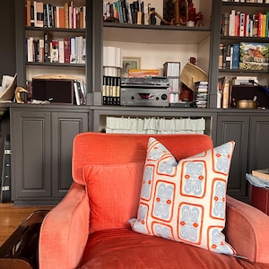 May include: An orange armchair with two pillows. One pillow is orange, and the other is cream-colored with a blue and orange floral pattern. A dark gray bookshelf filled with books is in the background.