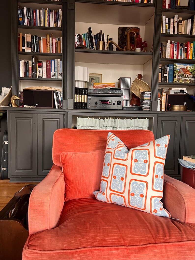 May include: An orange armchair with two pillows. One pillow is orange, and the other is cream-colored with a blue and orange floral pattern. A dark gray bookshelf filled with books is in the background.
