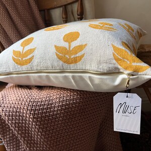 Puede incluir: Coj&iacute;n decorativo color crema con un estampado floral amarillo mostaza. El coj&iacute;n tiene una cremallera de lat&oacute;n y una etiqueta que dice "MUSE textiles hand printed home goods."