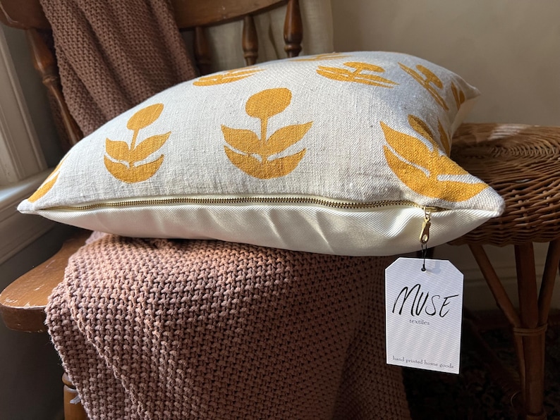 Puede incluir: Coj&iacute;n decorativo color crema con un estampado floral amarillo mostaza. El coj&iacute;n tiene una cremallera de lat&oacute;n y una etiqueta que dice "MUSE textiles hand printed home goods."