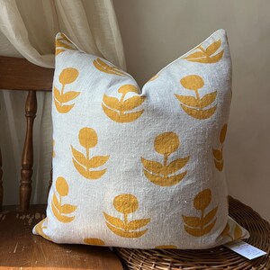 Puede incluir: Un coj&iacute;n decorativo cuadrado con un fondo gris claro y un patr&oacute;n repetido de dise&ntilde;os florales amarillos. El coj&iacute;n est&aacute; colocado sobre una silla de madera con un asiento tejido, mostrando una mezcla de texturas y colores.