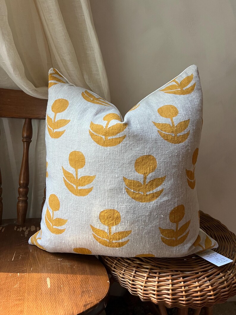 Puede incluir: Un coj&iacute;n decorativo cuadrado con un fondo gris claro y un patr&oacute;n repetido de dise&ntilde;os florales amarillos. El coj&iacute;n est&aacute; colocado sobre una silla de madera con un asiento tejido, mostrando una mezcla de texturas y colores.