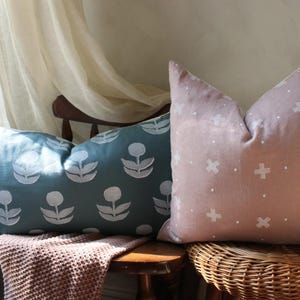 May include: Two decorative throw pillows. One is teal with a white floral pattern, the other is blush pink with a white cross pattern. They are placed on a wooden chair and a wicker stool.