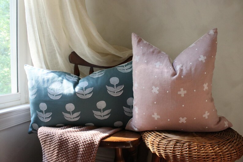 May include: Two decorative throw pillows. One is teal with a white floral pattern, the other is blush pink with a white cross pattern. They are placed on a wooden chair and a wicker stool.
