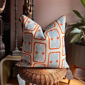 May include: Decorative throw pillow with a repeating pattern of light blue floral designs framed by orange borders on an off-white background. The pillow is placed on a woven wicker stool. A tag that says "MUSE" is attached to the pillow.
