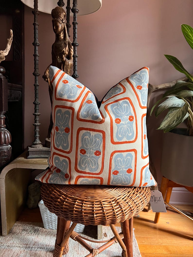 May include: Decorative throw pillow with a repeating pattern of light blue floral designs framed by orange borders on an off-white background. The pillow is placed on a woven wicker stool. A tag that says "MUSE" is attached to the pillow.