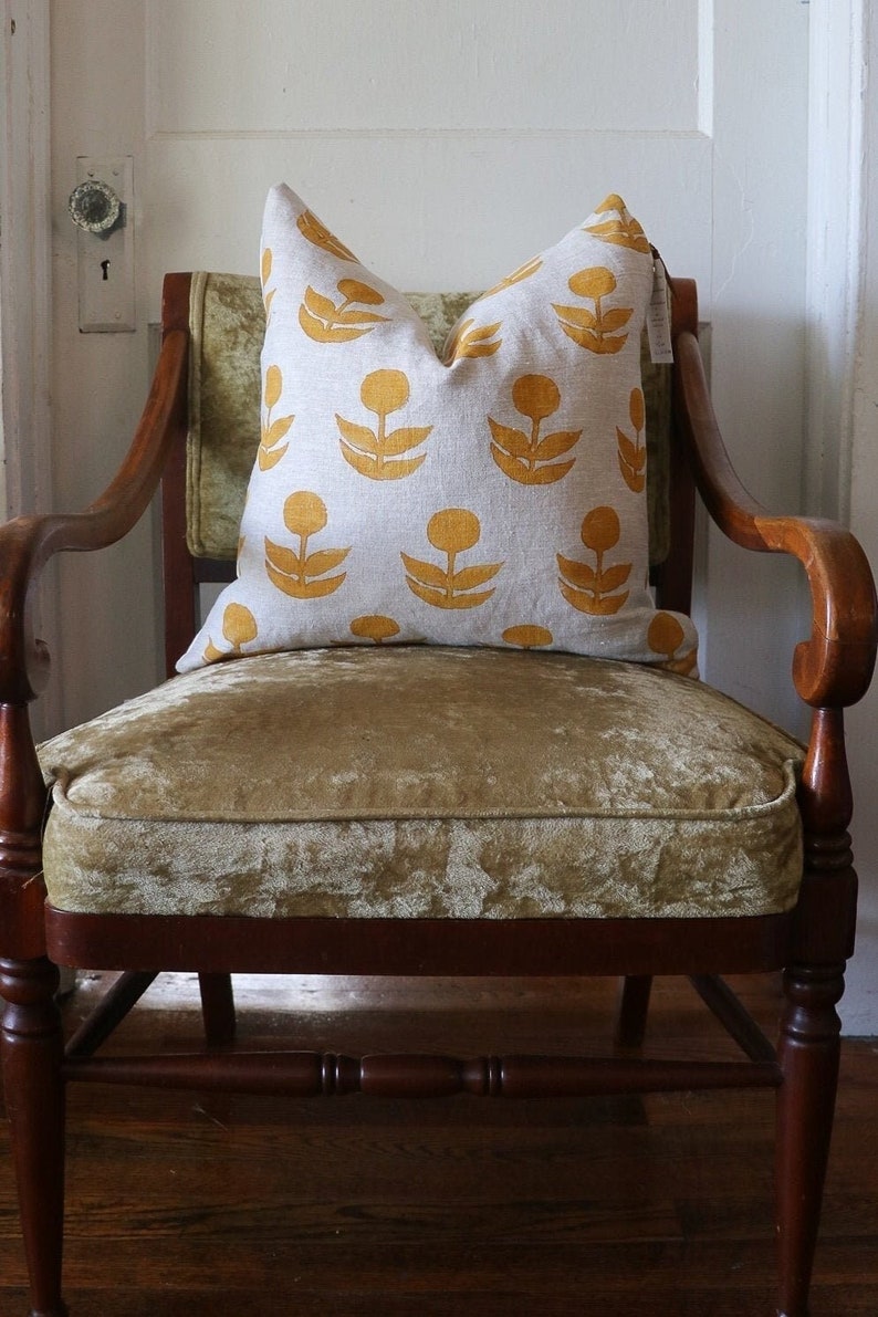 Puede incluir: Un coj&iacute;n decorativo con un estampado floral amarillo sobre un fondo gris claro descansa sobre una silla vintage. La silla tiene un marco de madera oscura y un coj&iacute;n de asiento de terciopelo dorado. El coj&iacute;n a&ntilde;ade color y patr&oacute;n.