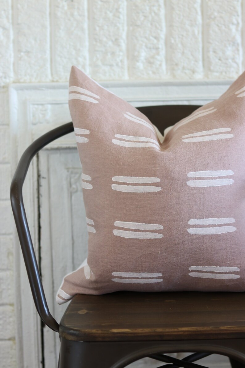 May include: A blush-colored decorative pillow with a repeating pattern of white horizontal stripes. The pillow is placed on a dark brown metal chair, offering a touch of modern design to any living space.