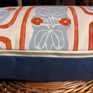 May include: Decorative pillow with a blue, white, and orange floral pattern. The pillow has a navy blue base and a brass zipper. The pillow is resting on a woven basket.