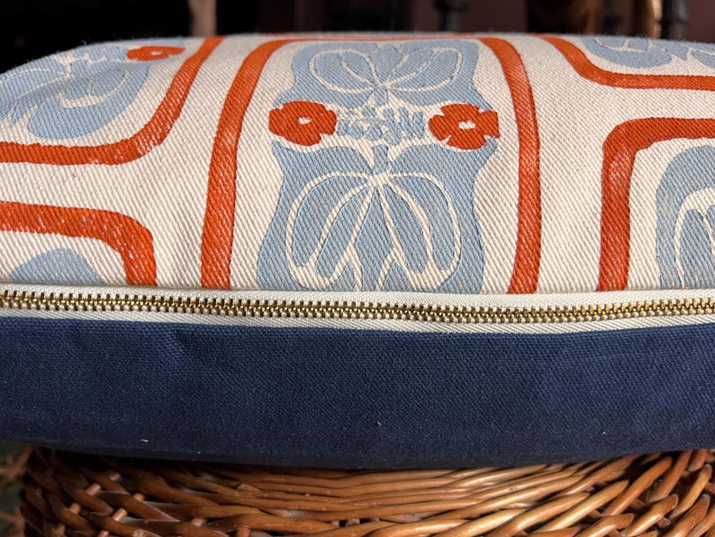 May include: Decorative pillow with a blue, white, and orange floral pattern. The pillow has a navy blue base and a brass zipper. The pillow is resting on a woven basket.