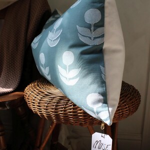 May include: A decorative pillow with a teal floral pattern and a white back. The pillow is propped on a wicker stool. A tag that says "MUSE textiles" is attached to the pillow.