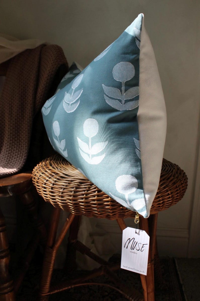 May include: A decorative pillow with a teal floral pattern and a white back. The pillow is propped on a wicker stool. A tag that says "MUSE textiles" is attached to the pillow.