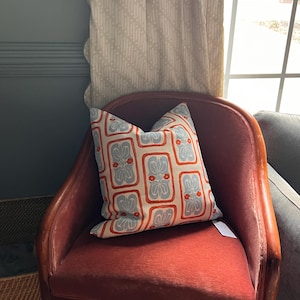 May include: A decorative pillow with a repeating pattern of blue and red floral designs on a cream background. The pillow is placed on a red upholstered chair with wooden arms. The chair is positioned near a window with patterned curtains.