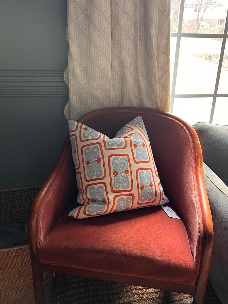 May include: A decorative pillow with a repeating pattern of blue and red floral designs on a cream background. The pillow is placed on a red upholstered chair with wooden arms. The chair is positioned near a window with patterned curtains.