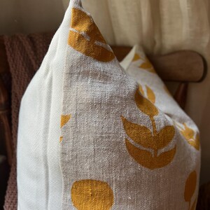 Puede incluir: Un coj&iacute;n decorativo con tela de lino beige y un patr&oacute;n repetido de dise&ntilde;os florales de color amarillo mostaza. El coj&iacute;n tiene un panel lateral a rayas y est&aacute; colocado en una silla de madera.